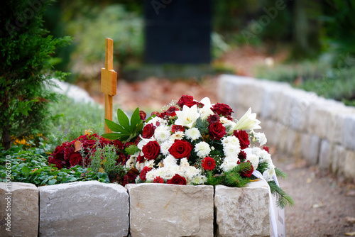 Funeral flowers of red roses and white dahlias and lilies with white ribbon on an urn grave in a modern funeral garden at a burial