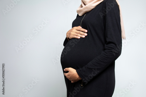 Pregnancy Concept side view. Unidentified pregnant woman wearing black dress touching her belly, muslim woman hijab, isolated on white
