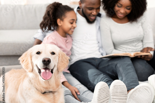 Leisure Time Concept. Portrait of cheerful black woman, man and girl reading paper book together and sitting on the floor in the blurred background, selective focus on happy golden retiever photo