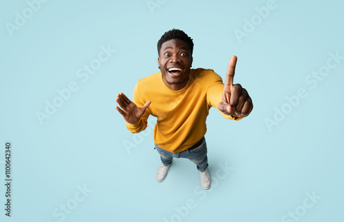 Top view of inspired creative african american millennial guy in casual outfit pointing finger up over blue studio background, having nice idea, showing eureka gesture, full length, copy space