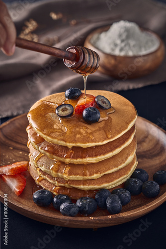 A stack of fluffy pancakes topped with strawberries, blueberries, and drizzled with honey. The image captures the moment honey is being poured, creating a perfect depiction of a delicious and comforti photo