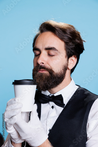 Classy doorkeeper smells coffee aroma on camera, wearing gloves and tie against blue background. Young man acting like a bellhop savouring smell of hot drink in cup, hotel employee. photo