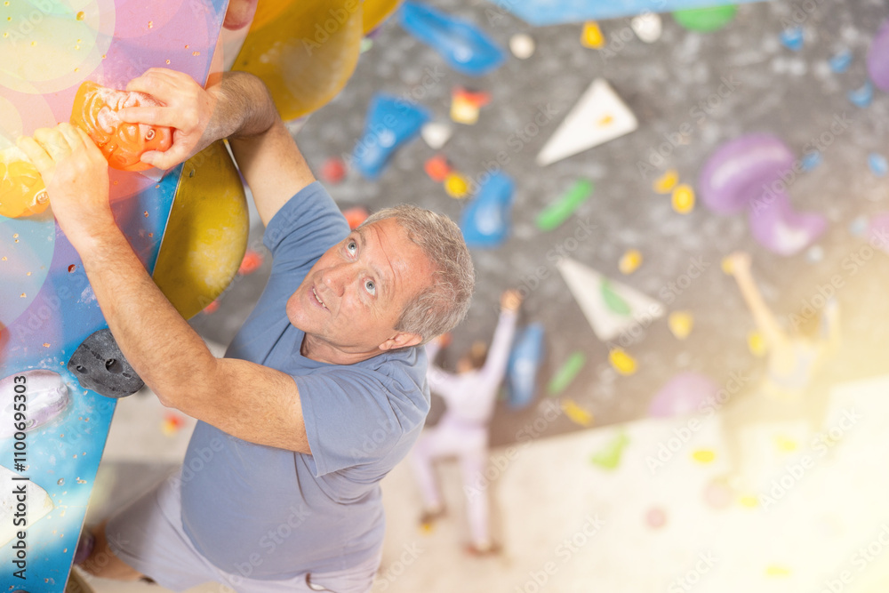Fototapeta premium Concentrated elderly man moving carefully on bouldering wall clinging to colorful hand and footholds. Popular activity in indoor climbing gym