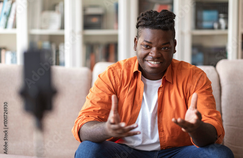 Blogging concept. Young African American chuby guy blogger in casual clothes streaming video online or recording tutorial for social media network, looking at the mobile phone on a tripod and smiling photo