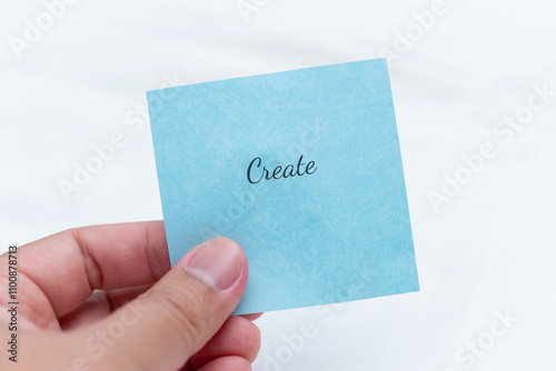 Hand holding blue note with text 'create' inspirational message minimalist setting close-up view for motivation photo