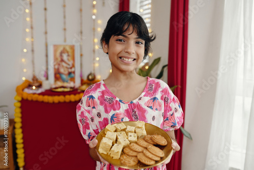 Young girl smiling while holding plate with traditional Indian snacks in festive setting. Background includes decorations and a religious figurine photo