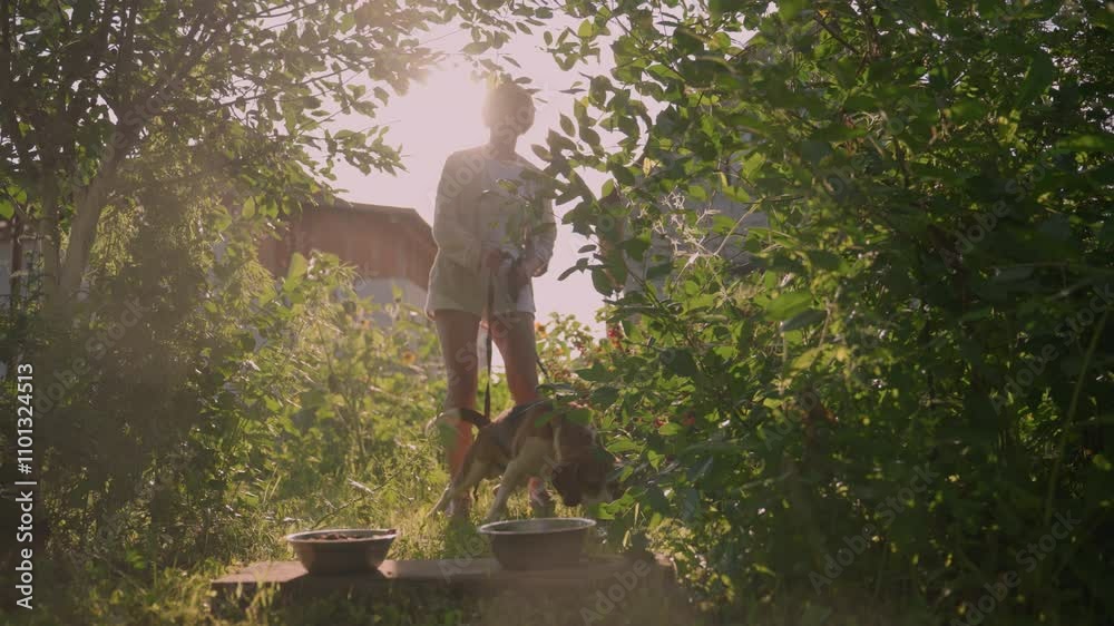 Dog owner standing outdoors holding leash as her dog attempts to move forward, she gently pulls it back, with two metal bowls placed on wooden plank in front of them, surrounded by greenery