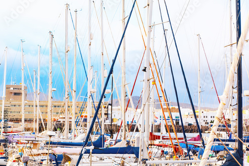 Densely packed marina with numerous sailboats docked closely together. Masts of the sailboats create a complex and visually striking pattern against the backdrop of buildings and a cloudy sky