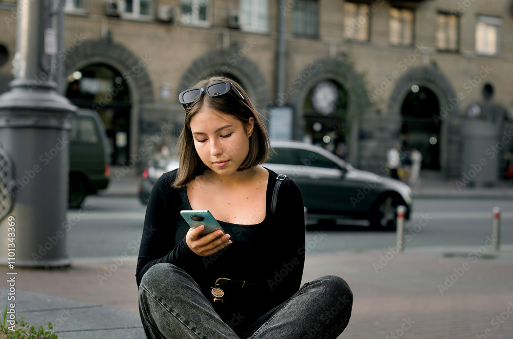 Fototapeta premium A young girl casually sits outdoors in an urban environment, fully engaged with her smartphone.