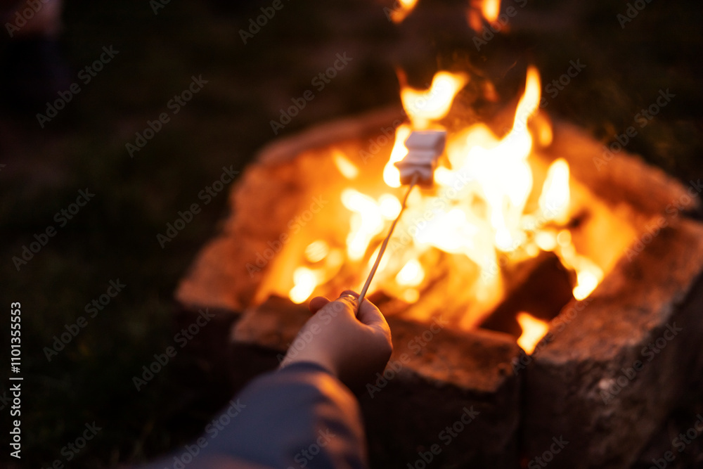 Fototapeta premium Close up of child's hand holding marshmallow on stick and baking it on campfire flame at night.