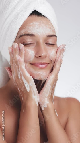 A serene woman wrapped in a towel gently washes her face with foaming cleanser during her skincare routine. Ideal for concepts of beauty, hygiene, self-care, and healthy skin.