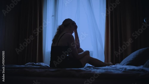 black silhouette upset unhappy woman sitting on bed dark room, cry wipes away tears with hand mood depression sadness emotion. Sad lonely Girl feels bad after party. dusk window curtains white fabric photo