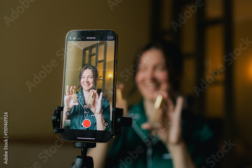 Plus size woman applying makeup on face with brush while recording vlog on smartphone for social media. Multiethnic fashion makeup artist vlogger live streaming makeup tutorial. Part of a series. UGC