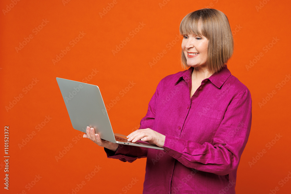 Elderly smiling happy blonde woman 50s years old she wears purple shirt casual clothes hold use work on laptop pc computer isolated on plain red orange background studio portrait. Lifestyle concept. Elderly smiling happy blonde woman 50s years old she wears purple shirt casual clothes hold use work on laptop pc computer isolated on plain red orange background studio portrait. Lifestyle concept.