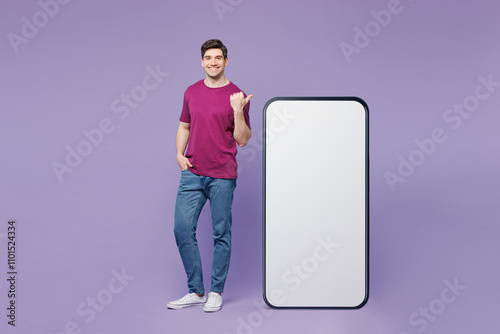 Full body young man he wear violet t-shirt casual clothes point finger on big huge blank screen mobile cell phone smartphone with workspace copy space mockup area isolated on plain purple background. photo