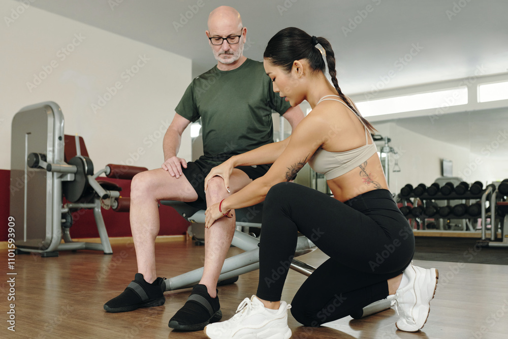 Female fitness trainer helping elderly man with leg exercise on gym floor. They are surrounded by various workout equipment and weights