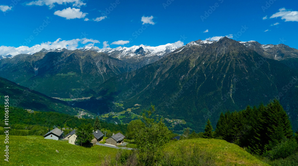 Fototapeta premium A charming village with quaint homes surrounded by vibrant green meadows and majestic mountain peaks under a bright blue sky, evoking peace and tranquility.