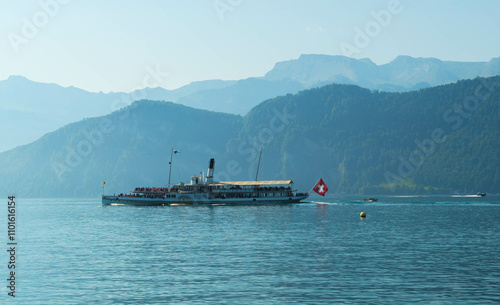 A vintage paddle steamer boat sails across a calm lake with Swiss mountains in the background, featuring a Swiss flag and a buoy toward a serene landscape.