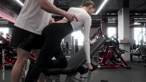In the gym, a supportive male personal trainer provides guidance to a woman as she lifts a dumbbell. Athletic Support Male Personal Trainer Assisting Woman in Weightlifting
