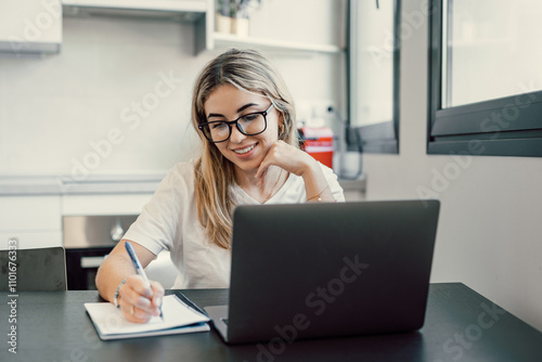 Millennial blonde girl sit at desk in living room study on laptop making notes, concentrated young woman work on computer write in notebook, take online course or training at home, education concept.