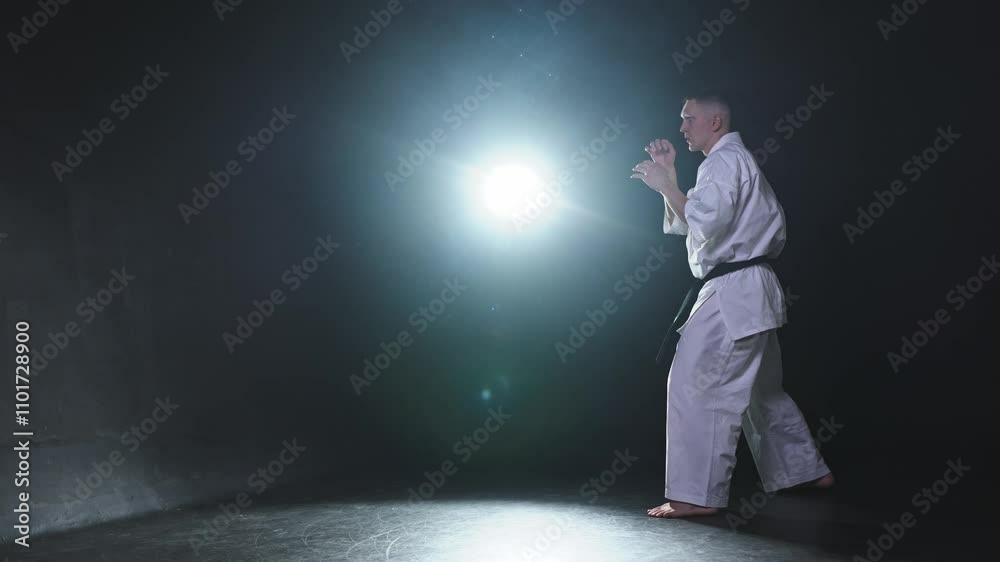Strong man fighter in white kimono for karate martial arts with black belt preparing for fight training in black background. Man training moves kicks practicing shadow fight ready for sparring Strong man fighter in white kimono for karate martial arts with black belt preparing for fight training in black background. Man training moves kicks practicing shadow fight ready for sparring