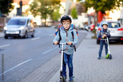 Two school kid boys in safety helmet riding with scooter in the city with backpack on sunny day. Happy children in colorful clothes biking on way to school.