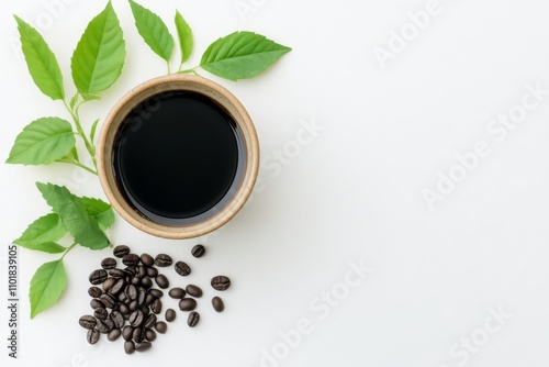 Dark coffee in a bowl accompanied by fresh leaves and coffee beans on a white...