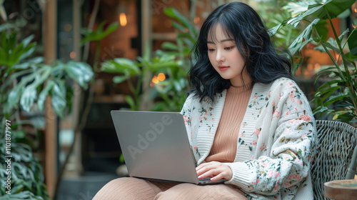 Beautiful Korean girl operating an unbranded laptop in an outdoor cafe, black hair, plump figure, real person, wearing fashion clothes, sitting, cafe-style aesthetic plant background.