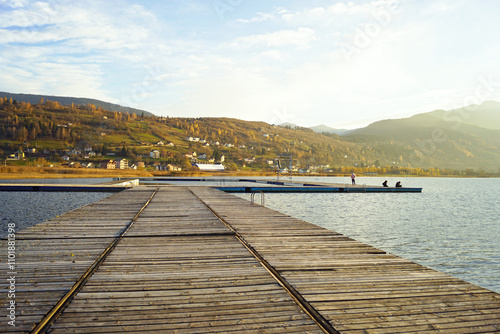 Outdoor recreation: a long wooden pier and people relaxing on it against the backdrop of the town of Plav illuminated by the golden rays of the setting sun. Landscape from Lake Plav in Montenegro. photo