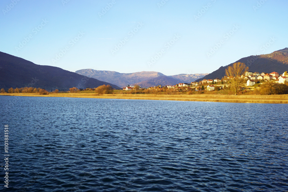 Landscape from the town of Plav: bright blue waters of the lake, thickets of golden reeds, trees with autumn leaves, houses and mountain slopes. Travel to the east of Montenegro: recreation in nature. Landscape from the town of Plav: bright blue waters of the lake, thickets of golden reeds, trees with autumn leaves, houses and mountain slopes. Travel to the east of Montenegro: recreation in nature.