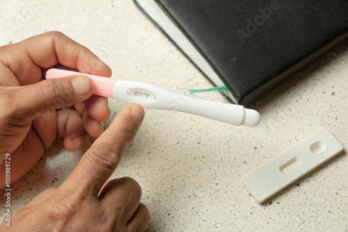 a woman's hand holding a pregnancy test kit on a granite table and books background. The test kit results show two lines, which means positive pregnancy. photo