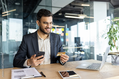 Businessman in office holds smartphone and credit card, completing online transaction. Smiling professional multitasks with phone, laptop, expressing confidence. Modern workspace reflects digital age