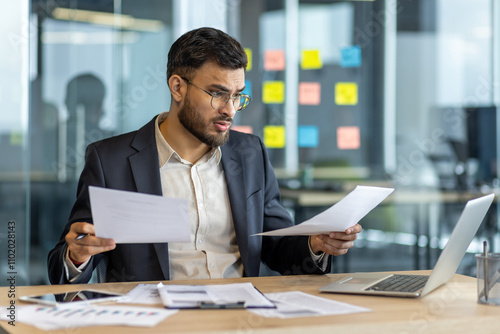 Businessman sitting at office desk with confused expression, analyzing important documents. Professional holds paperwork in hands, attempting to comprehend reports. Corporate setting insight.