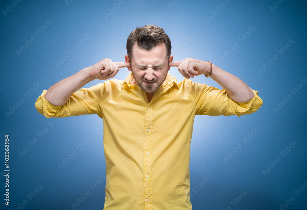 Noise - loud. Waist-up portrait of a young angry unhappy stressed man covering his ears closing eyes over blue background, dresses in yellow shirt Noise - loud. Waist-up portrait of a young angry unhappy stressed man covering his ears closing eyes over blue background, dresses in yellow shirt
