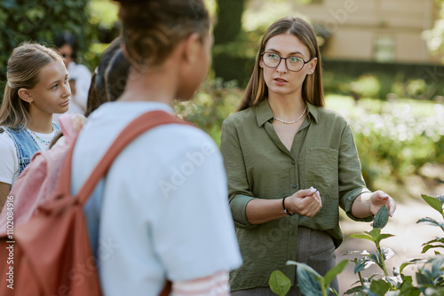 Young female teacher wearing eyeglasses speaking about plants during outdoor class in botanical garden