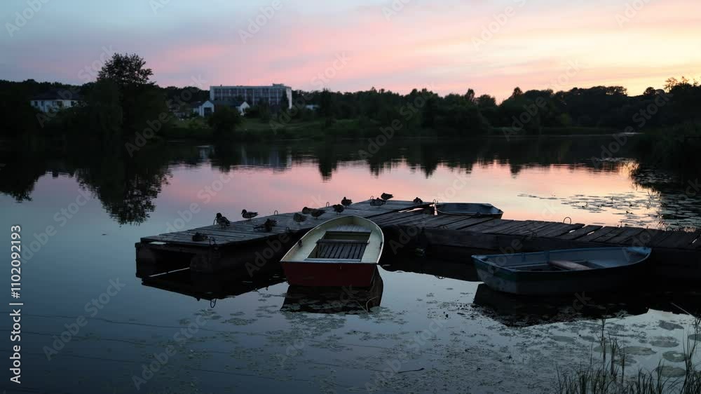 Beautiful sunset on the river, lake. Multi-colored boats sway on the water at the pier. An ancient wooden pier. A flock of ducks is resting. The red sun is reflected in the river. Green trees. Beautiful sunset on the river, lake. Multi-colored boats sway on the water at the pier. An ancient wooden pier. A flock of ducks is resting. The red sun is reflected in the river. Green trees.