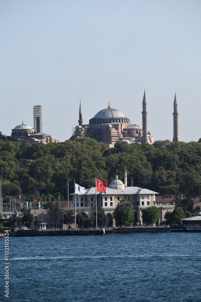 Obraz premium View of Hagia Sophia with Turkish and Yeşilay flags in the foreground along the Bosphorus in Istanbul, framed by lush greenery and the waterfront on a sunny day 
