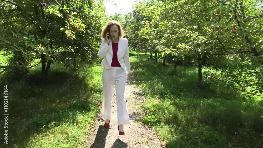 Woman walking along apple tree alley and talking on mobile phone, gesturing, front view Woman walking along apple tree alley and talking on mobile phone, gesturing, front view