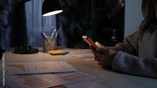 Foto Adult business woman sitting at desk working late at night in dark office, watching scrolling social media on smartphone, procrastinating work