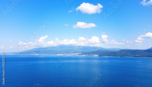 Serene coastal landscape with blue sea, mountains, and fluffy clouds under a bright sky.