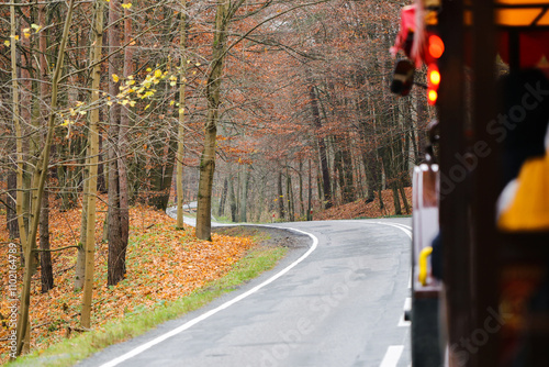 Tractor driving through an autumn forest on a scenic asphalt road. Tourist ride with wagons carrying passengers. Winding path in a landscape park. Golden yellow leaves all around.