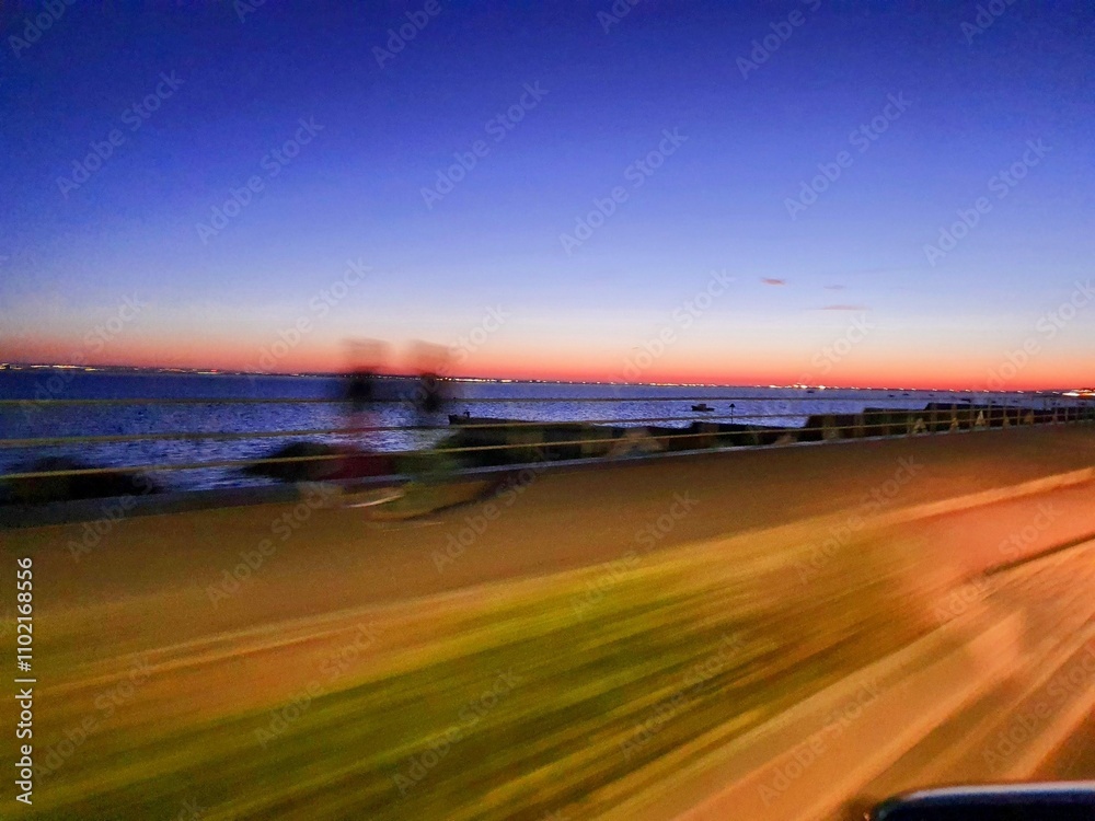 Naklejka premium Sunset Along the Waterfront Road With Blurred Figures and Tranquil Water