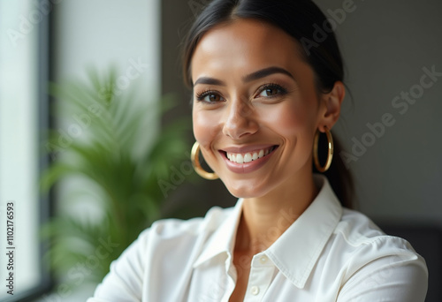 A smiling woman with golden hoop earrings  wearing a white shirt  near a window.