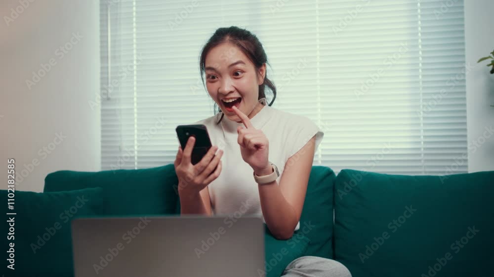 Freelancer is working from home, sitting on the sofa and using her laptop, when she receives great news on her smartphone and starts laughing
