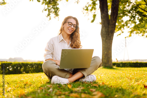 Happy woman sitting on a green clearing with a laptop. Freelancer, education online. Communication online.