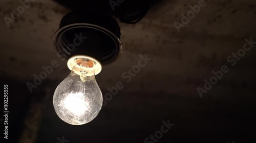 A close-up on a black background shows an incandescent lamp that turns on and off, creating a flickering effect. The filament, emitting a warm light, seems to dance in the wind. A light bulb in a dark