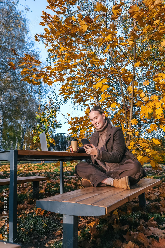 Fototapeta premium woman working remotely in a beautiful autumn park, sitting on a bench with a laptop. The serene outdoor environment is perfect for remote work or study, surrounded by fall foliage.