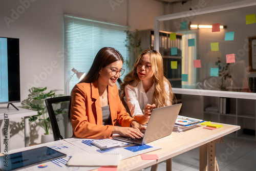 Two businesswomen working late on laptop in office with sticky notes on glass partition wall behind