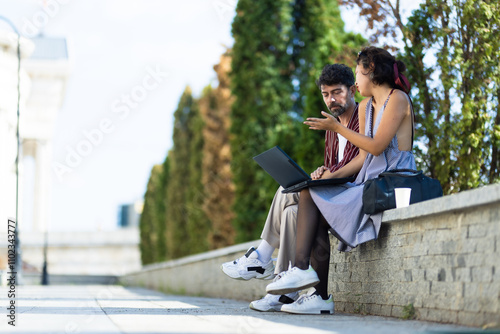 Two colleagues discussing a project while sitting on an outdoor park bench with a laptop and coffee, engaging in a productive work meeting