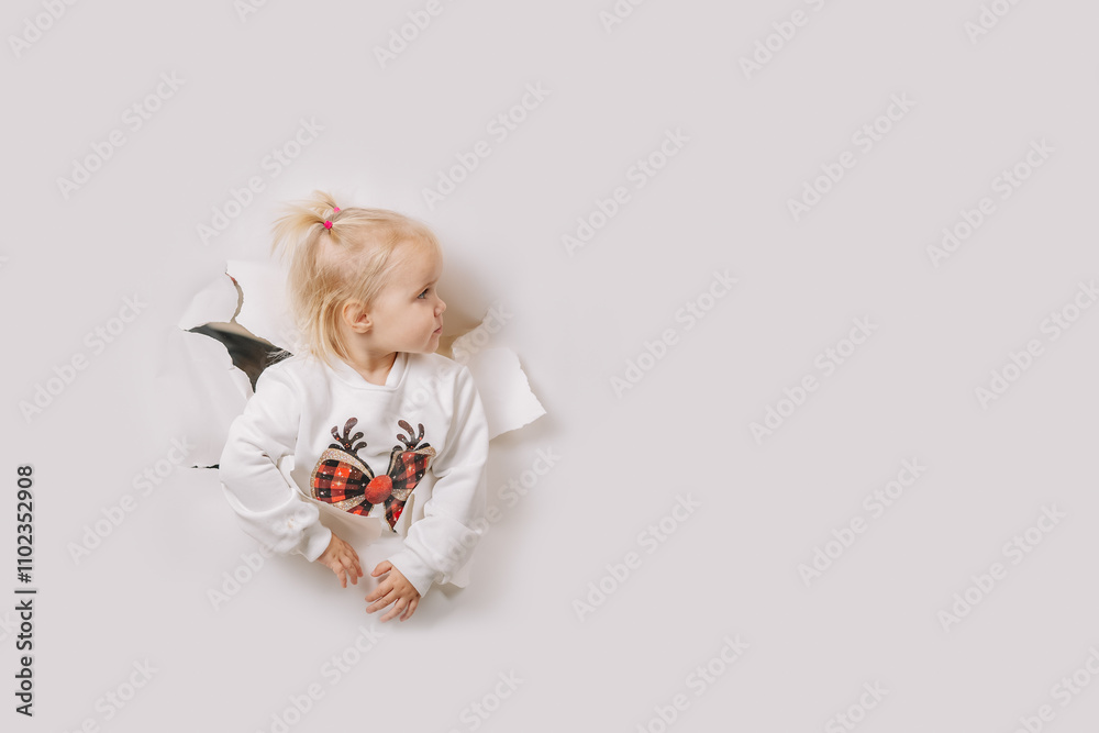 A toddler girl in a festive reindeer sweater looks sideways, emerging through a torn white paper background. Minimalistic design with copy space. A toddler girl in a festive reindeer sweater looks sideways, emerging through a torn white paper background. Minimalistic design with copy space.